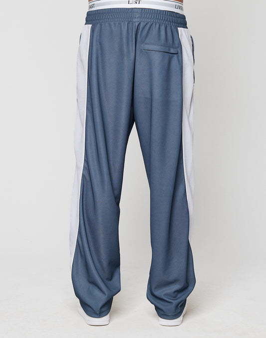 The lower half of a person is shown wearing LIVE FAST DIE YOUNG’s LFDY DY Trackpants—blue, wide fit track pants with a white side stripe and elastic waistband—against a plain white background.