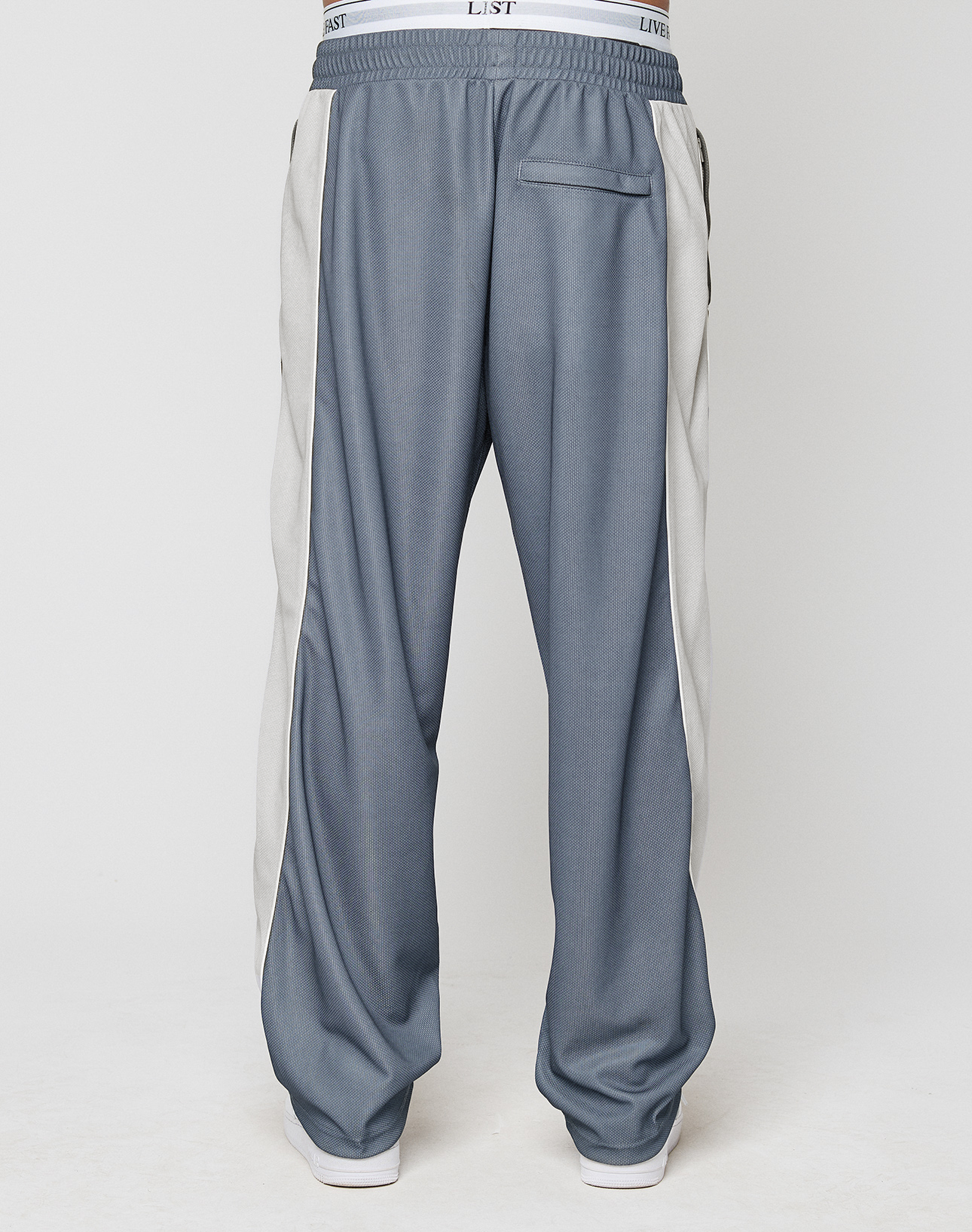 A person is shown from the back wearing LIVE FAST DIE YOUNG LFDY DY Trackpants in a wide fit blue design with light gray side panels and a visible white elastic waistband, standing against a plain light background.