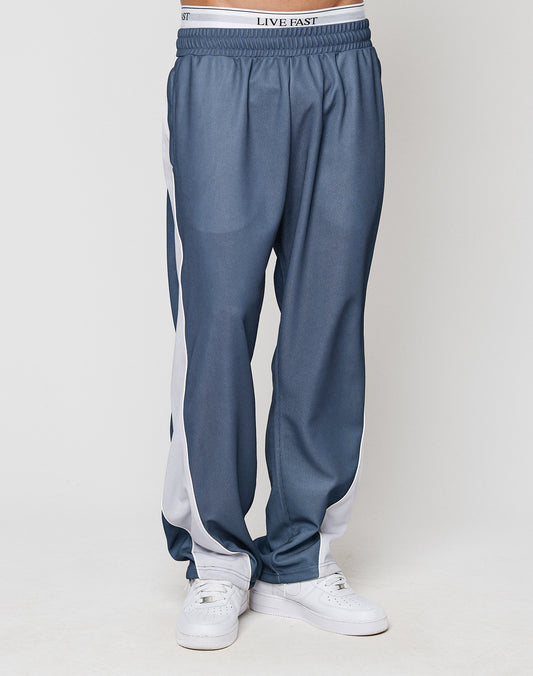 A person wears LIVE FAST DIE YOUNG LFDY DY Trackpants in wide fit blue with white side stripes and elastic waistband reading LIVE FAST, paired with white sneakers, standing against a plain light background.