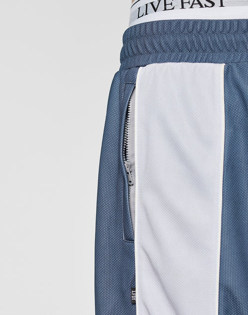 Close-up of a person wearing LFDY DY Trackpants by LIVE FAST DIE YOUNG, in wide fit blue and white polyester, featuring a zipper pocket and elastic waistband. The LIVE FAST waistband of their underwear is slightly visible above the pants.