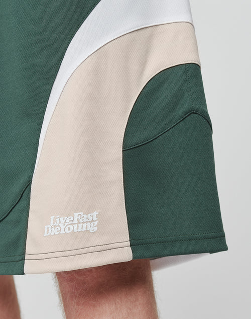 A close-up of green, beige, and white Basic Trackshorts by LIVE FAST DIE YOUNG with an oversized fit and elastic waistband. The brands Live Fast Die Young logo is printed in white near the hem. Part of a leg is visible below the shorts.