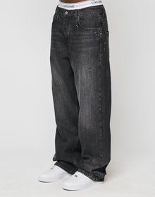 Someone wears the LFDY Baggy Denim by LIVE FAST DIE YOUNG—black jeans with wide straight legs, light fades, unique color splatters, subtle distressed details—paired with white sneakers and a visible LIVE FAST waistband. | model