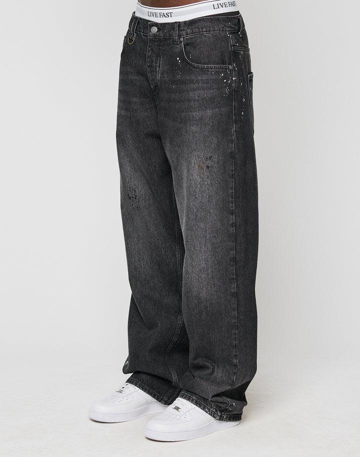 Someone wears the LFDY Baggy Denim by LIVE FAST DIE YOUNG—black jeans with wide straight legs, light fades, unique color splatters, subtle distressed details—paired with white sneakers and a visible LIVE FAST waistband. | model