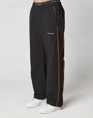 A person in LIVE FAST DIE YOUNGs Live Fast Trackpants—wide fit, black with orange piping, elastic waist with drawstring, and LIVE FAST on the thigh—paired with white sneakers, stands against a plain light background.