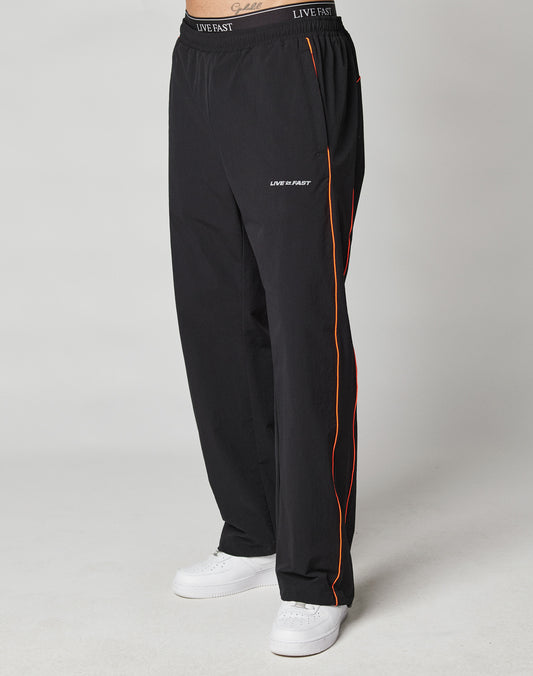 A person wears LIVE FAST DIE YOUNGs black Live Fast Trackpants with orange piping, LIVE FAST text, reflective logo print on the left thigh, elastic waistband with drawcord, and white sneakers against a plain light background.