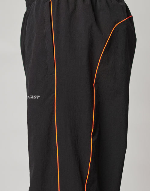 The LIVE FAST DIE YOUNG Live Fast Trackpants are black with an elastic waistband and drawstring, thin orange side piping, a reflective logo print on the thigh, and a wide fit, photographed against a plain background.