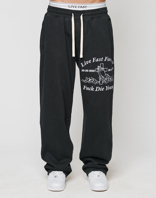 A person wears LFDY Praying Jogger sweatpants by LIVE FAST DIE YOUNG, featuring an elastic waistband with white drawstrings, white sneakers, and oversized fit with Live Fast Forever text and graphic details.