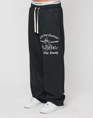 A person wearing LIVE FAST DIE YOUNGs LFDY Praying Jogger—black baggy sweatpants with white graphics, an oversized fit, and elastic waistband—paired with white sneakers. Only the lower body is visible.