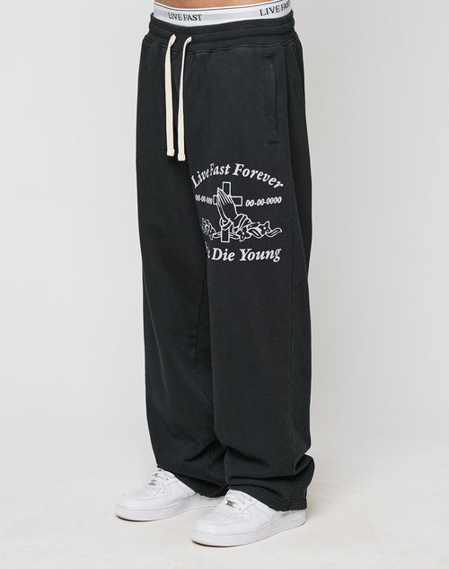 A person wearing LIVE FAST DIE YOUNGs LFDY Praying Jogger—black baggy sweatpants with white graphics, an oversized fit, and elastic waistband—paired with white sneakers. Only the lower body is visible.