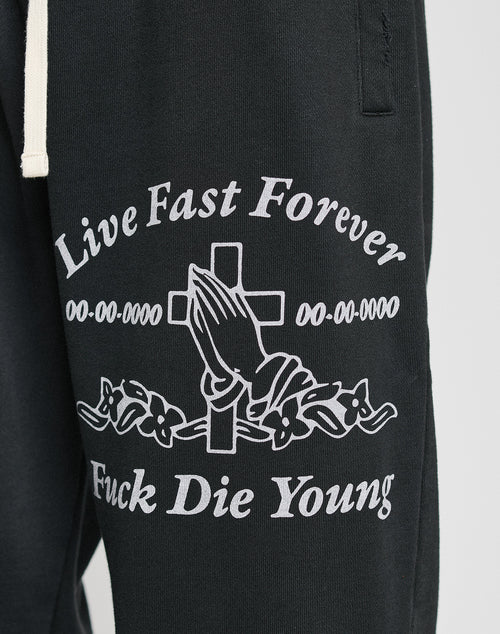 The LFDY Praying Jogger by LIVE FAST DIE YOUNG features black sweatpants with an elastic waistband, white praying hands graphic in front of a cross, “Live Fast Forever” & “Fuck Die Young” text, floral accents, and a relaxed oversize fit.