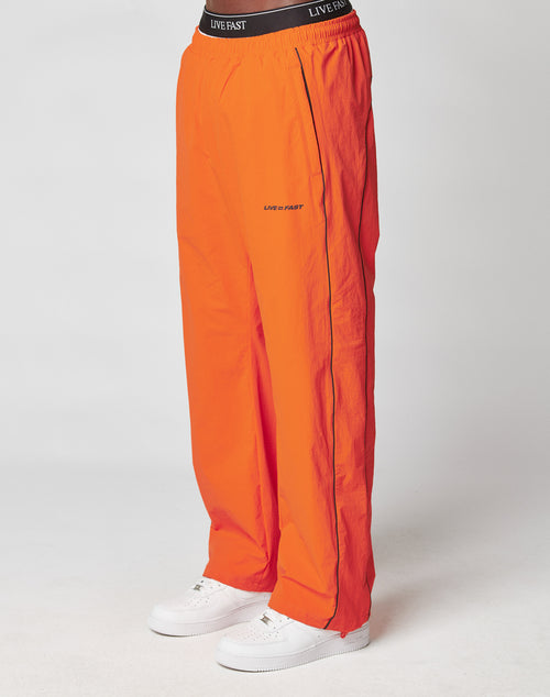 A person wears LIVE FAST DIE YOUNG’s Live Fast Trackpants in bright orange with black piping, a wide elastic waistband featuring LIVE FAST, and a reflective logo print, paired with white sneakers against a plain background.