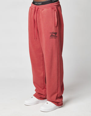 A person wears the LIVE FAST DIE YOUNG Live Fast Sweatsuit Jogger—loose-fitting red pants with a vintage wash, elastic waistband reading Live Fast, and thigh logo—paired with white sneakers, against a plain white background.
