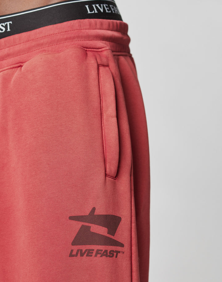 Close-up of a person in LIVE FAST DIE YOUNG Live Fast Sweatsuit Jogger—red, vintage-wash joggers with deep pockets and a black LIVE FAST logo near the hip. Black underwear waistband with LIVE FAST text peeks above the elastic waist.