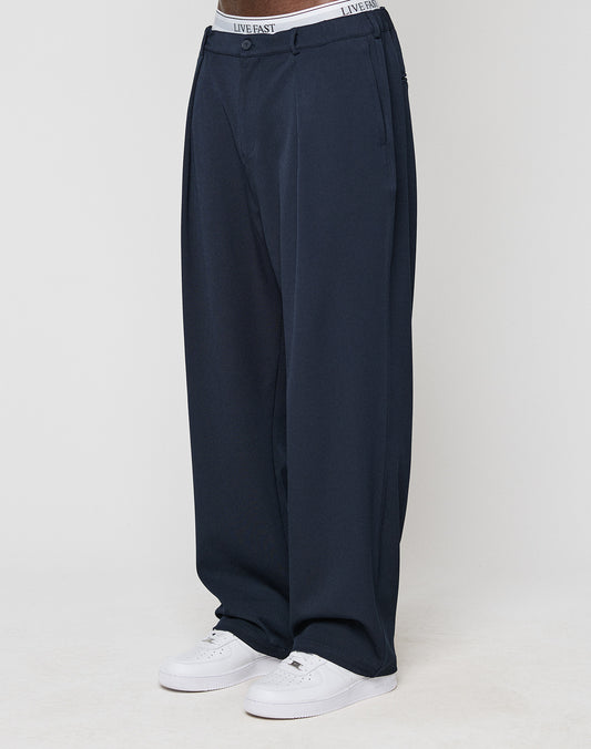 Someone stands against a white background wearing LIVE FAST DIE YOUNG Wide Casual Pants in navy blue with an elastic waistband, paired with white sneakers and a visible white waistband that says LIVE FAST.
