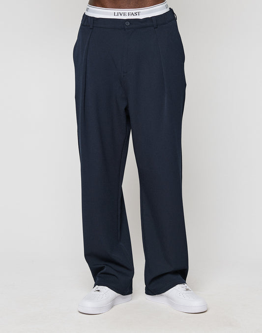 A person wears dark blue Wide Casual Pants by LIVE FAST DIE YOUNG with a gerades Bein and wide fit, styled with white sneakers. The LIVE FAST elastischer Bund waistband is visible above the pants. The background is plain white.