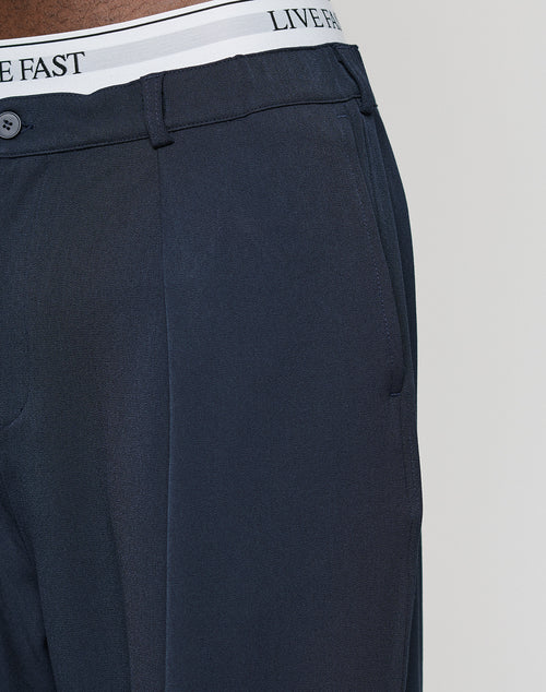 Close-up of a person wearing LIVE FAST DIE YOUNG Wide Casual Pants in dark navy with a straight leg and wide fit. The pants have belt loops, a button, and reveal a white elastic waistband printed with “LIVE FAST.”.