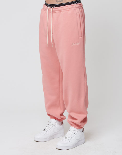 A person wearing LIVE FAST DIE YOUNG Loose Jogger in pink with elastic waistband and drawstrings, paired with white sneakers and a black waistband with white lettering, stands against a plain white background.
