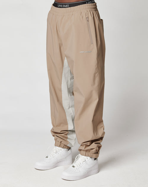 A person wears beige and light gray Basic Shell Pants by LIVE FAST DIE YOUNG, featuring reflective print and an elastic waistband with drawstring, paired with white sneakers, standing against a plain white background.