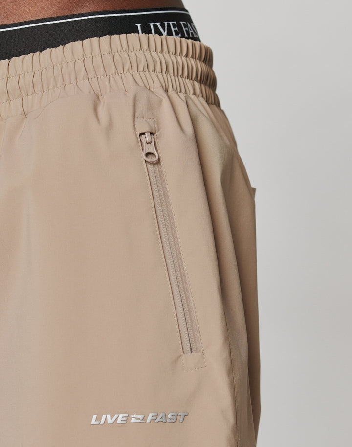 Close-up of beige Basic Shell Pants by LIVE FAST DIE YOUNG featuring an elastic waistband with drawstring, zippered side pocket, and reflective LIVE FAST print logo near the hem. Black waistband with LIVE text also visible.