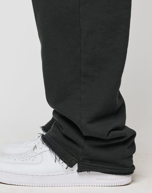 A close-up side view of a person wearing the LIVE FAST DIE YOUNG LFDY Lightweight Jogger in black, featuring frayed hems and an elastic waistband in an oversize fit over white sneakers, standing on a light surface.