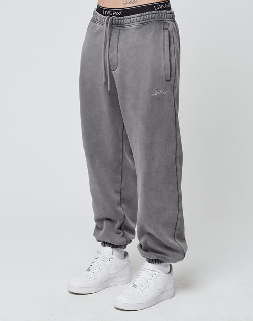 A person stands against a plain white background wearing LIVE FAST DIE YOUNG Loose Jogger sweatpants in grey, featuring elastic cuffs and white Live Fast branding on the waistband, paired with white Nike sneakers.