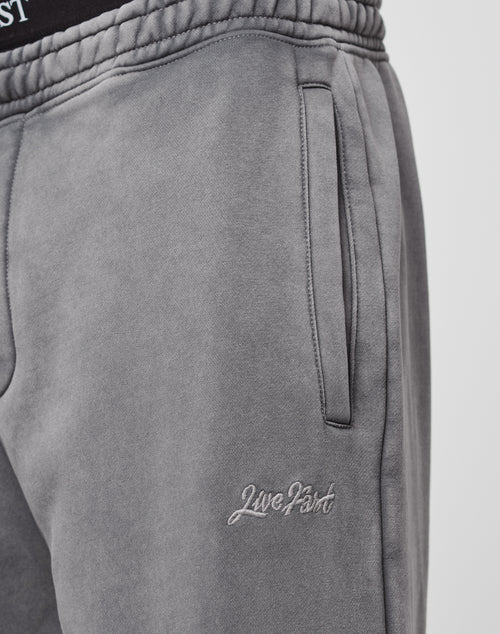 Close-up of a person wearing the Loose Jogger by LIVE FAST DIE YOUNG in grey, featuring an elastic waistband, side pocket, and “LiveFast” logo embroidered in white on the left thigh. Part of another waistband with text appears above the pants.