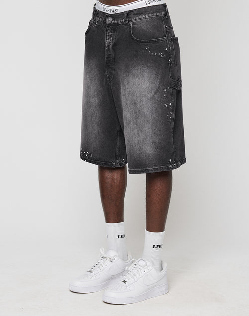 A person wearing LIVE FAST DIE YOUNG Carpenter Jorts—faded black, baggy knee-length denim shorts with weiße Farbspritzer—plus white LIVE FAST socks and white Nike sneakers, standing against a plain white background. 100% Baumwolle.
