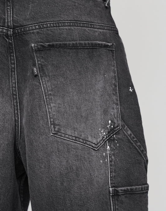 Close-up of the back pocket on LIVE FAST DIE YOUNG Carpenter Jorts in dark gray with white paint splatters by the seam, set against a plain light background. Baggy fit; made from 100% cotton.
