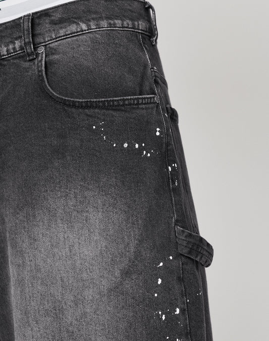 Close-up of LIVE FAST DIE YOUNG Carpenter Jorts in black, baggy fit, 100% cotton with white paint-splatter details and a side utility pocket, shown from waist to mid-thigh against a plain background.