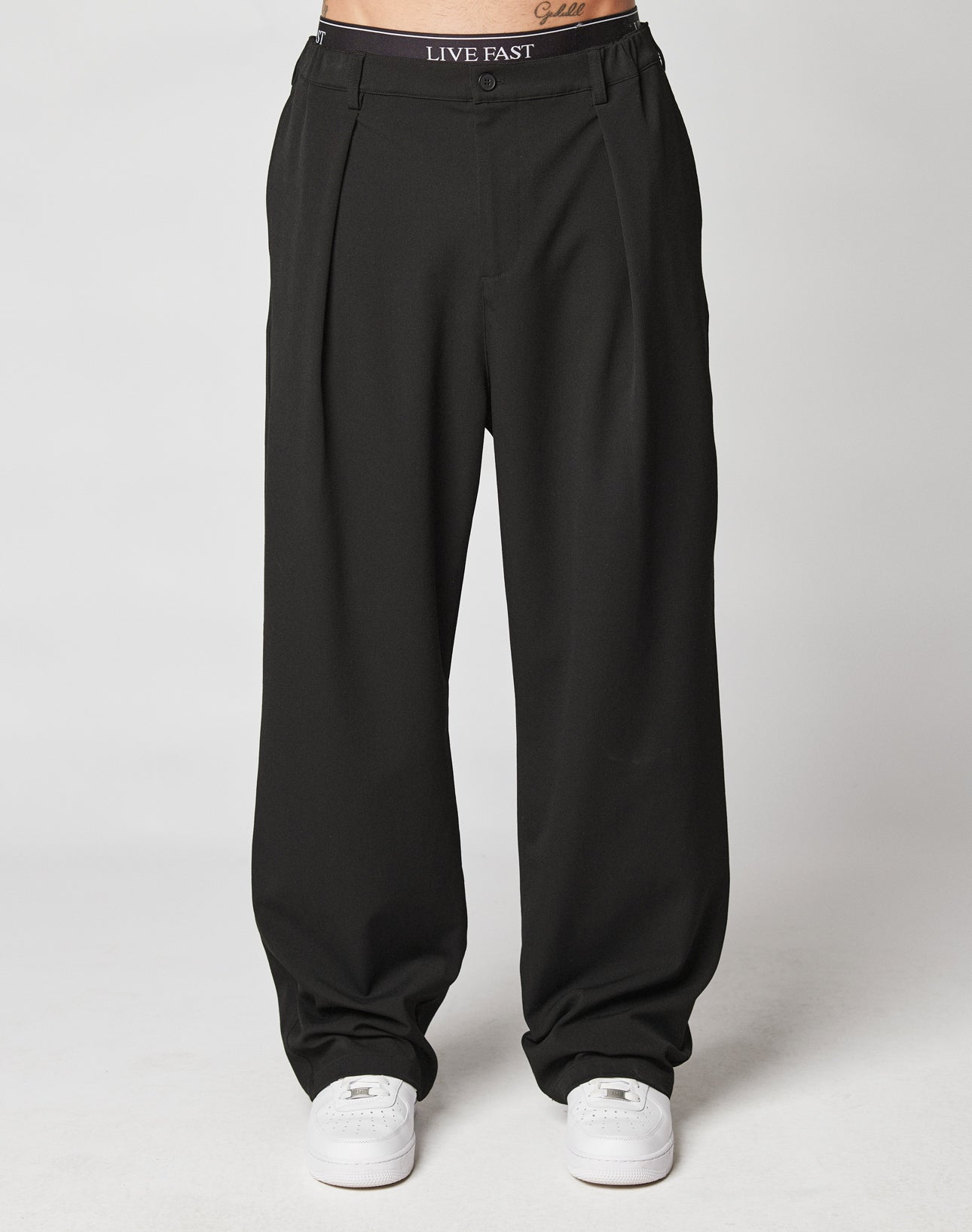 Someone stands against a plain white background wearing LIVE FAST DIE YOUNG Wide Casual Pants—loose black pleated trousers with a LIVE FAST waistband—paired with white sneakers.