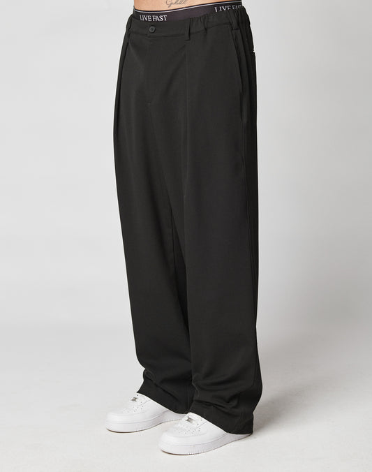 A person wears loose-fitting black Wide Casual Pants by LIVE FAST DIE YOUNG, featuring a printed waistband, paired with white sneakers. Only the lower body is shown against a plain light background.