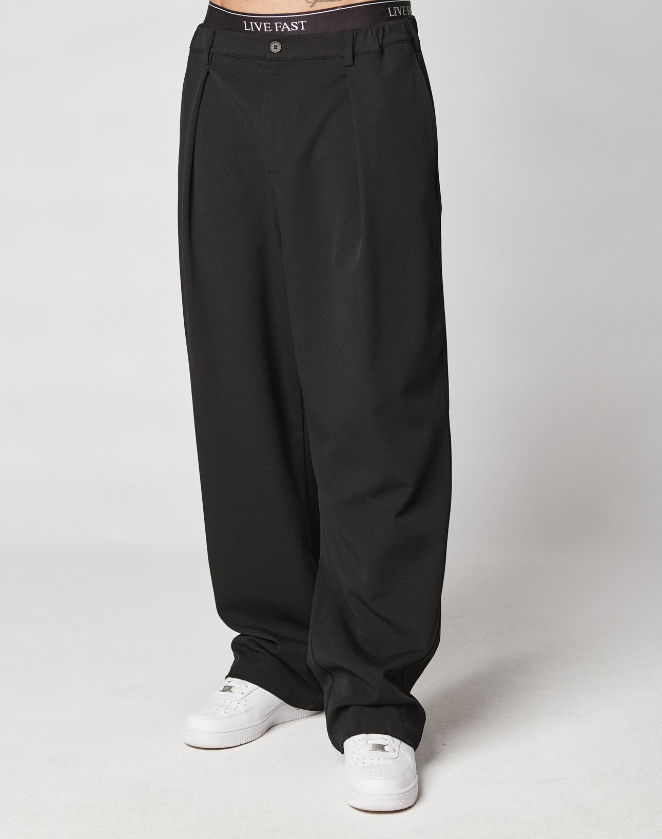 A person wears black LIVE FAST DIE YOUNG Wide Casual Pants with pleats, white sneakers, and a visible waistband reading LIVE EAST, standing against a plain light background.