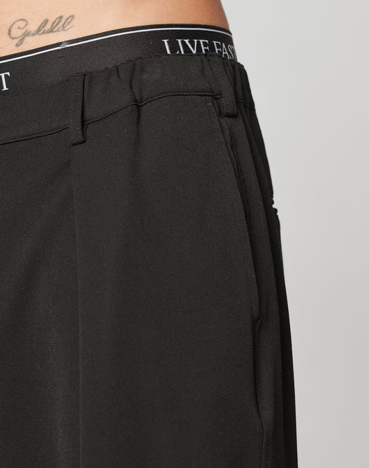 Close-up of a person wearing black Wide Casual Pants by LIVE FAST DIE YOUNG, featuring LIVE FAST on the waistband and a cursive Goodluck tattoo above the waist. Background is plain and light-colored.