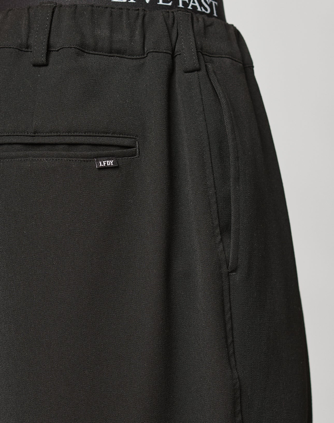 Close-up of LIVE FAST DIE YOUNG Wide Casual Pants in black, showing a back welt pocket with LFDY tag, elastic waistband, and "LIVE FAST" text on the lining. Perfect Herren Stoffhose for a relaxed look.