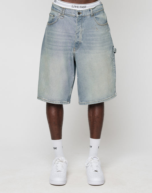 A person wears LIVE FAST DIE YOUNG Carpenter Jorts in a baggy fit, light blue, knee-length style with visible LIVE FAST waistband, paired with white sneakers and socks, standing against a plain white background.