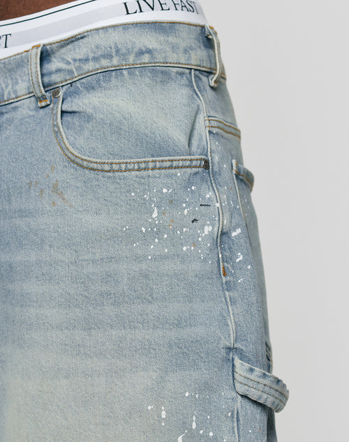 Close-up of LIVE FAST DIE YOUNG Carpenter Jorts in baggy light blue denim with white paint splatters and a utility pocket. The white LIVE FAST waistband of underwear is visible above these 100% cotton shorts.