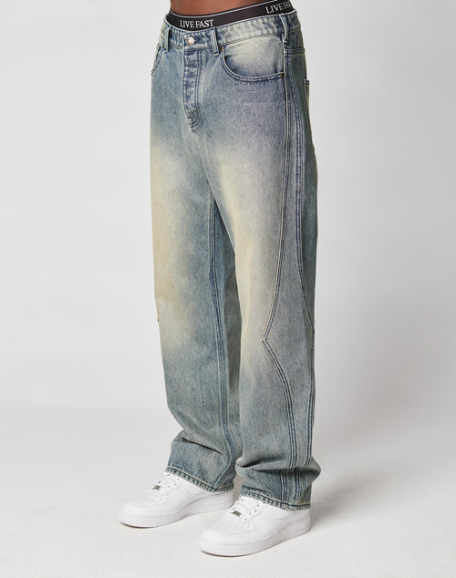 A person wears LIVE FAST DIE YOUNGs Rhomb Wide Denim—baggy, light-wash jeans with diamond-shaped panels and a faded yellow tint—paired with white sneakers and black LIVE FAST waistband underwear, photographed on a plain white background.