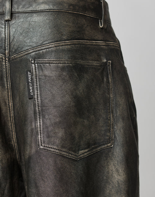 Close-up of the back pocket on black, distressed Baggy Leather Pants by LIVE FAST DIE YOUNG. A small black flag label with LIVE FAST is on the left side. The leather shows visible texture and fading.