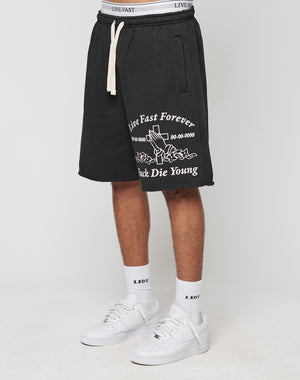 A person wears LFDY Praying Shorts by LIVE FAST DIE YOUNG—regular fit, black with white “Live Fast Forever, Fuck Die Young” text and rose graphic, elastic waistband, side pockets—paired with “LDY” socks and white sneakers.