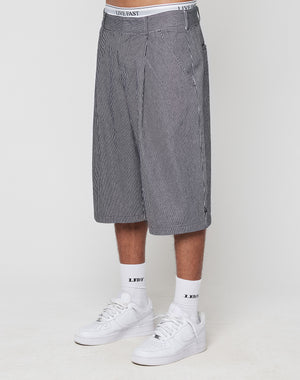 A person wears black and white pinstriped LFDY Twill Shorts by LIVE FAST DIE YOUNG with an oversize fit and white LIVE FAST waistband, paired with white L.E.D.Y socks and sneakers, standing against a plain light background.