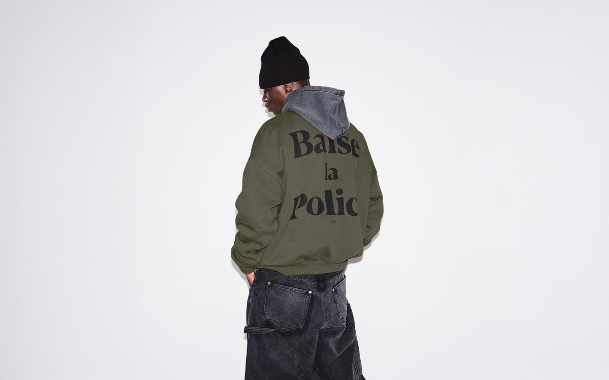 A person stands against a plain white background, wearing a black beanie, layered olive green jacket with the words Baisse la Police printed on the back, a gray hoodie, and loose black jeans.