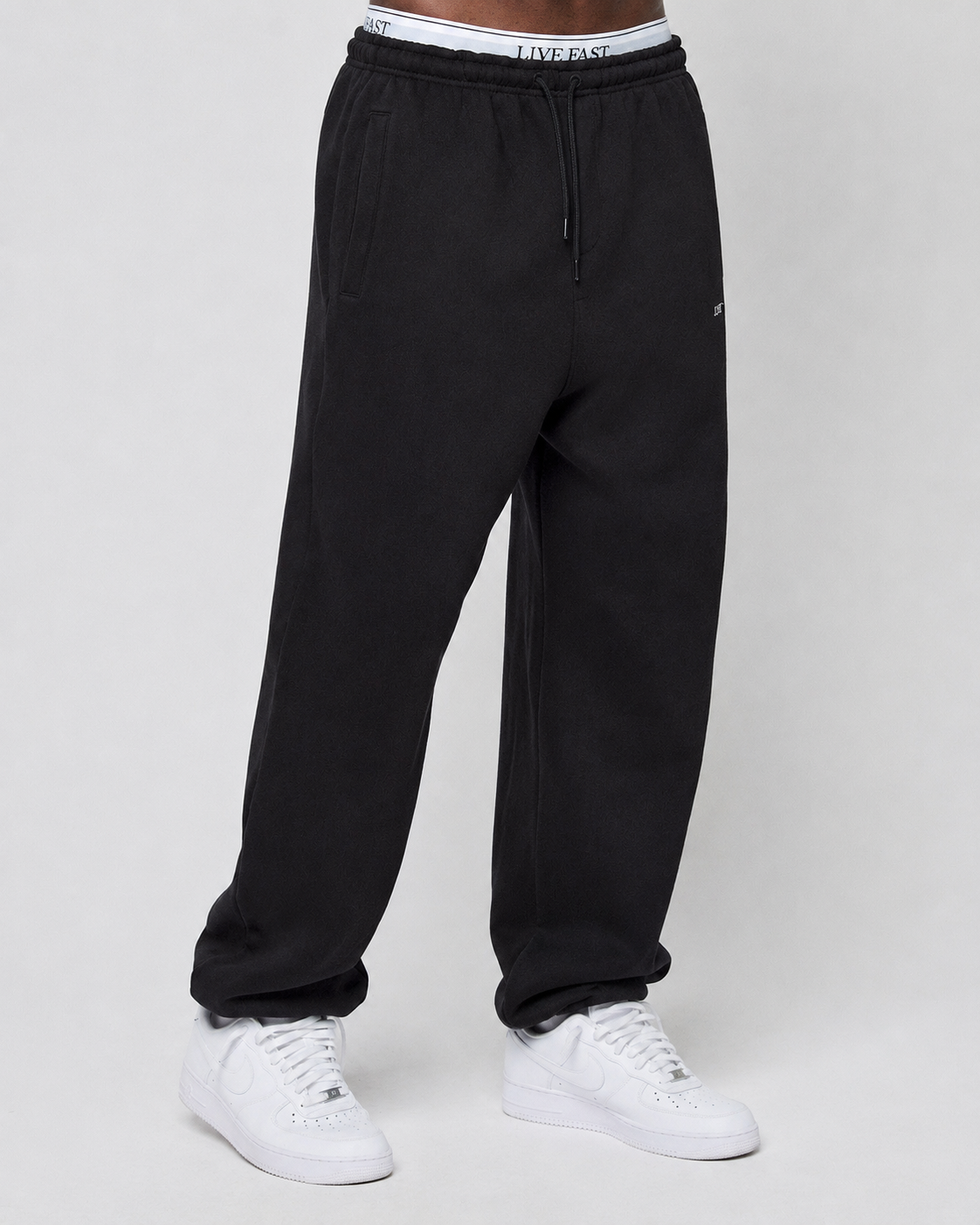 A person in a Basic Loose Jogger by LIVE FAST DIE YOUNG with side pockets, a white waistband reading LIVE FAST, and white Nike sneakers stands against a plain light background.