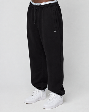 A person wears a Basic Loose Jogger by LIVE FAST DIE YOUNG with “LEFT” logo on the left leg, white high-top sneakers, and an elastic waistband featuring “LIVE FAST.” The background is plain and light-colored.