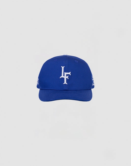 The LF 10th Snapback by LIVE FAST DIE YOUNG is a blue 100% cotton baseball cap with a white LF Multi-Logo embroidered on the front, shown on a light gray background. Features an adjustable snap closure.