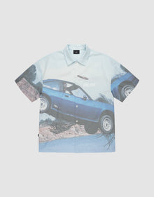The LFDY Crash Kid Shirt by LIVE FAST DIE YOUNG features short sleeves and an oversized blue car print mid-jump over dirt, with "LIVE FAST" and a bold logo on the car door. | plain