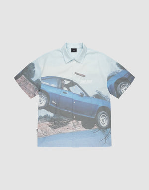 The LFDY Crash Kid Shirt by LIVE FAST DIE YOUNG features short sleeves and an oversized blue car print mid-jump over dirt, with "LIVE FAST" and a bold logo on the car door. | plain