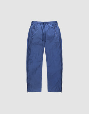 The LIVE FAST DIE YOUNG Zip Tape Track Pants are loose blue nylon pants with a crinkled texture, elastic waistband, side pockets, and ankle zippers, shown against a plain light background. | plain