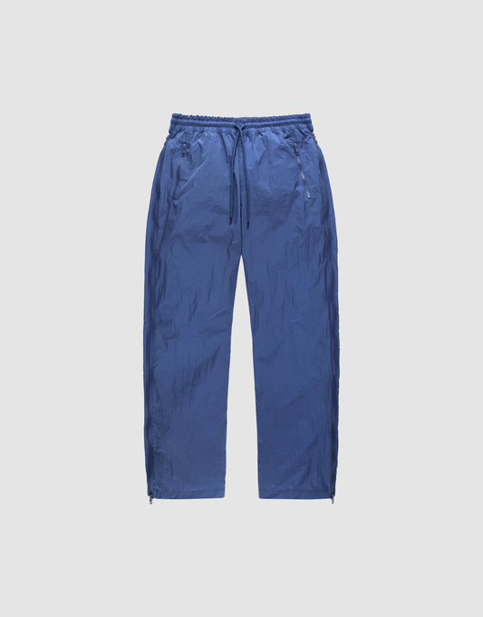 The LIVE FAST DIE YOUNG Zip Tape Track Pants are loose blue nylon pants with a crinkled texture, elastic waistband, side pockets, and ankle zippers, shown against a plain light background. | plain