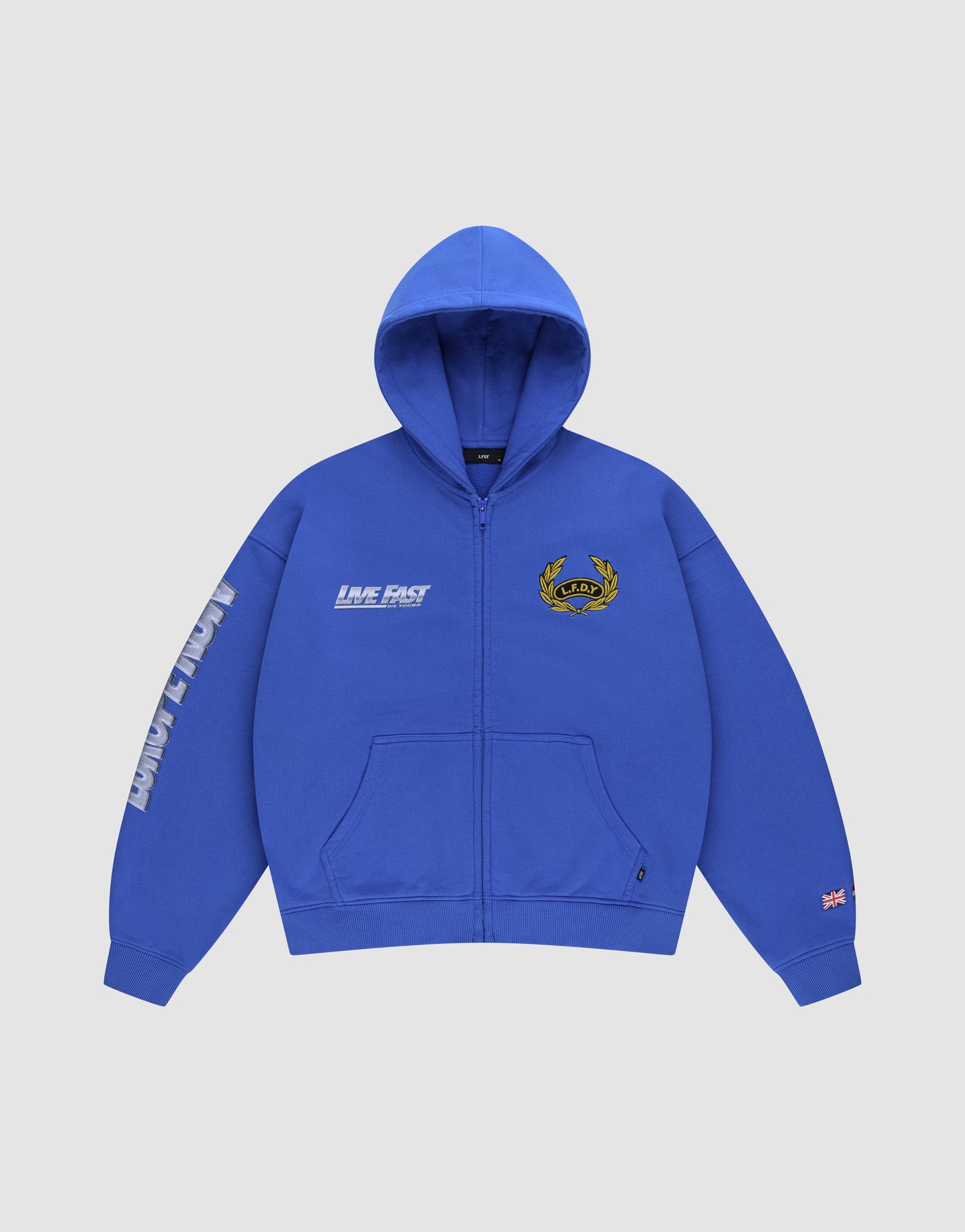 The LFDY Team Hooded Zip by LIVE FAST DIE YOUNG is a royal blue oversize fit zip-up hoodie with “LIVE FAST” on the chest, Multilogo embroidery including a laurel crest, “FAST LANE” on one sleeve, and a flag patch on the other.