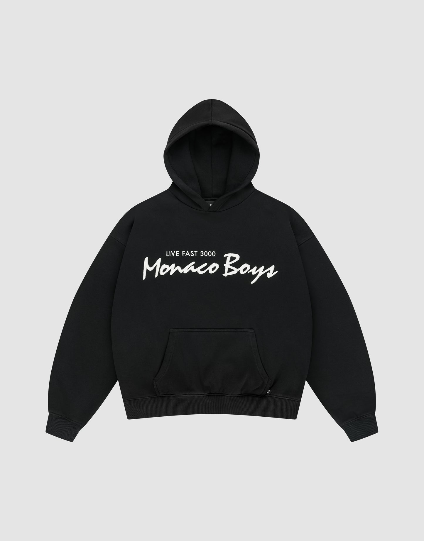 The Monaco Boys Hooded by LIVE FAST DIE YOUNG is a black oversized hoodie with a front pocket, featuring "LIVE FAST 3000 Monaco Boys" printed in white on the chest and finished with a subtle flag-label detail. | plain